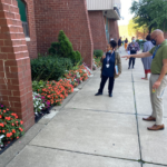 Judges stand on the sidewalk and look at flowers planted at foot of building